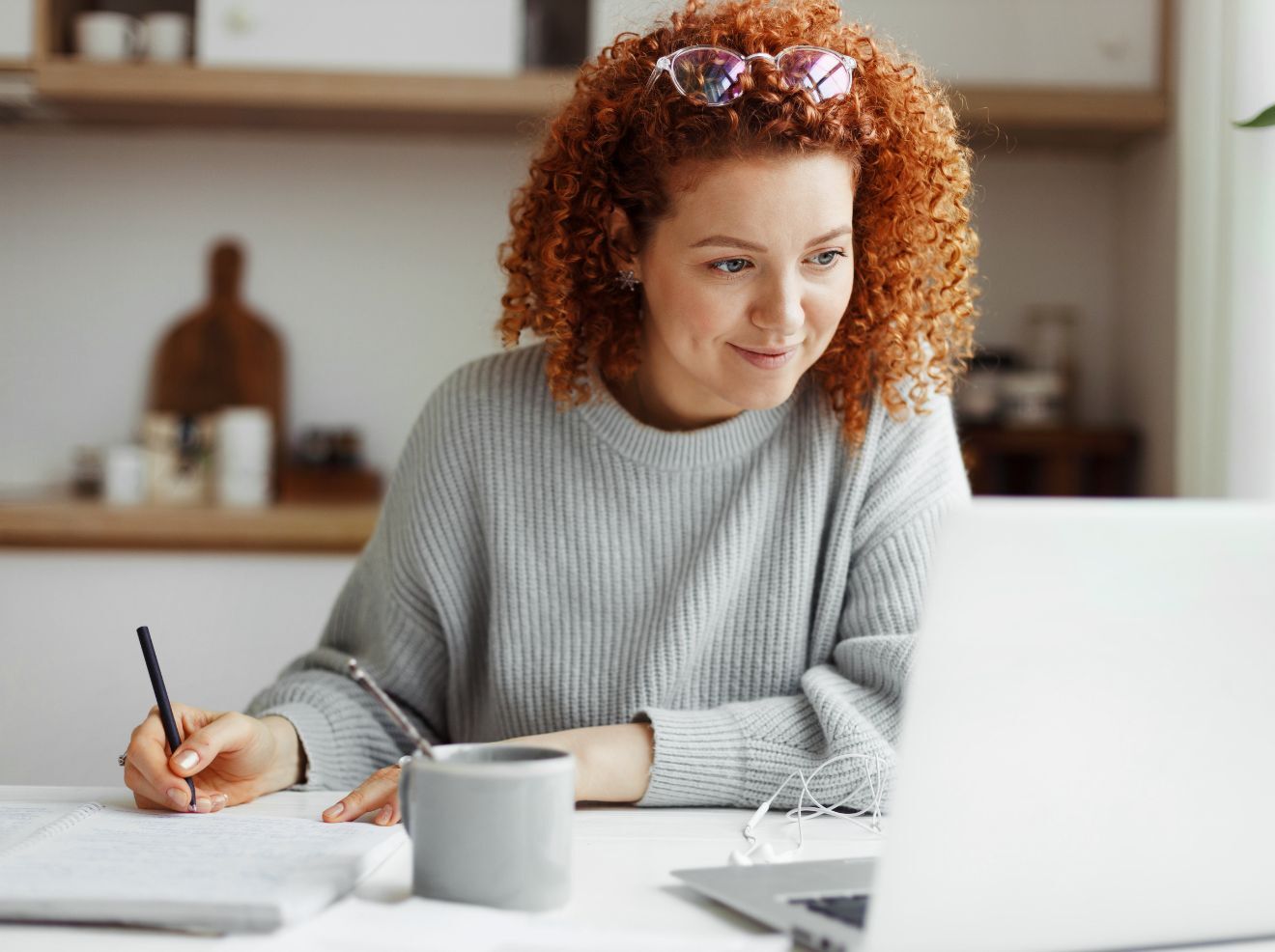 A woman with curly red hair writes in a notebook while looking at a laptop on a white desk, with a mug and earphones beside her.